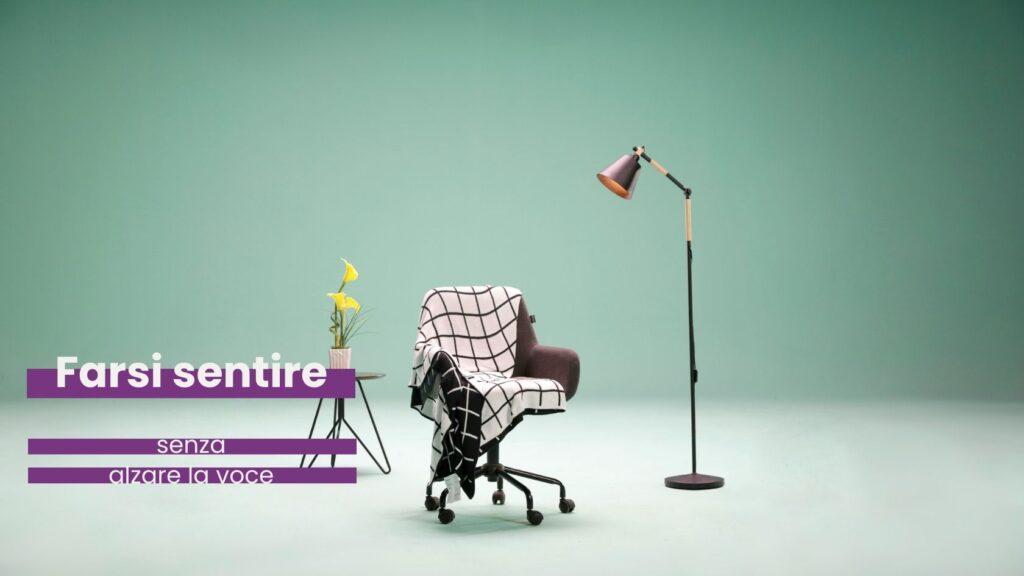 Una foto minimalista in cui ci sono tre oggetti. Da sinistra a destra: un tavolino di ferro con sopra un vasetto con due calle, una sedia da ufficio con un plaid a quadri, una lampada a stelo.